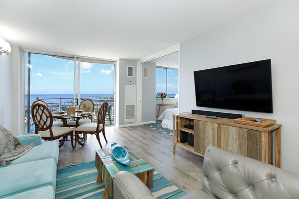 A modern living room with a large TV, comfortable seating, a dining area, and a balcony with a view of the ocean. - 11th Floor Waikiki Ocean, Lagoon & Marina Views! Walk to Beach! Washlet! (Honolulu)