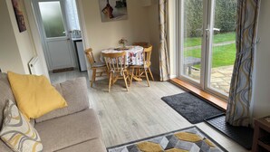 A living room with a sofa, a dining table with chairs, a rug, and a view of a garden through a glass door. - Holiday Let Annex; Self Contained; Sleeps 2; One minute from beach (Nr CHICHESTER)