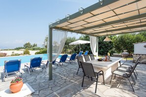 An outdoor patio with a pool, blue lounge chairs, a table with chairs, and a pergola with a canopy.