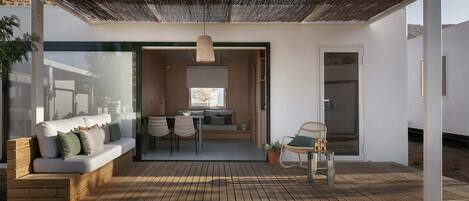 A modern outdoor living space with a wooden deck, a sofa with cushions, a dining table with chairs, and a hanging light fixture.