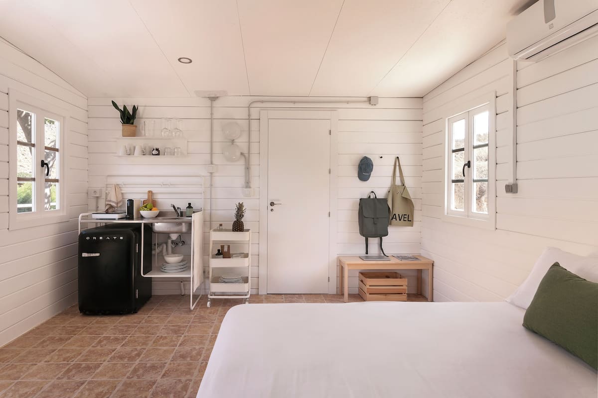 A room with a bed, a desk, a small table, a refrigerator, a shelf with glassware, and a door with a towel hanging on it.