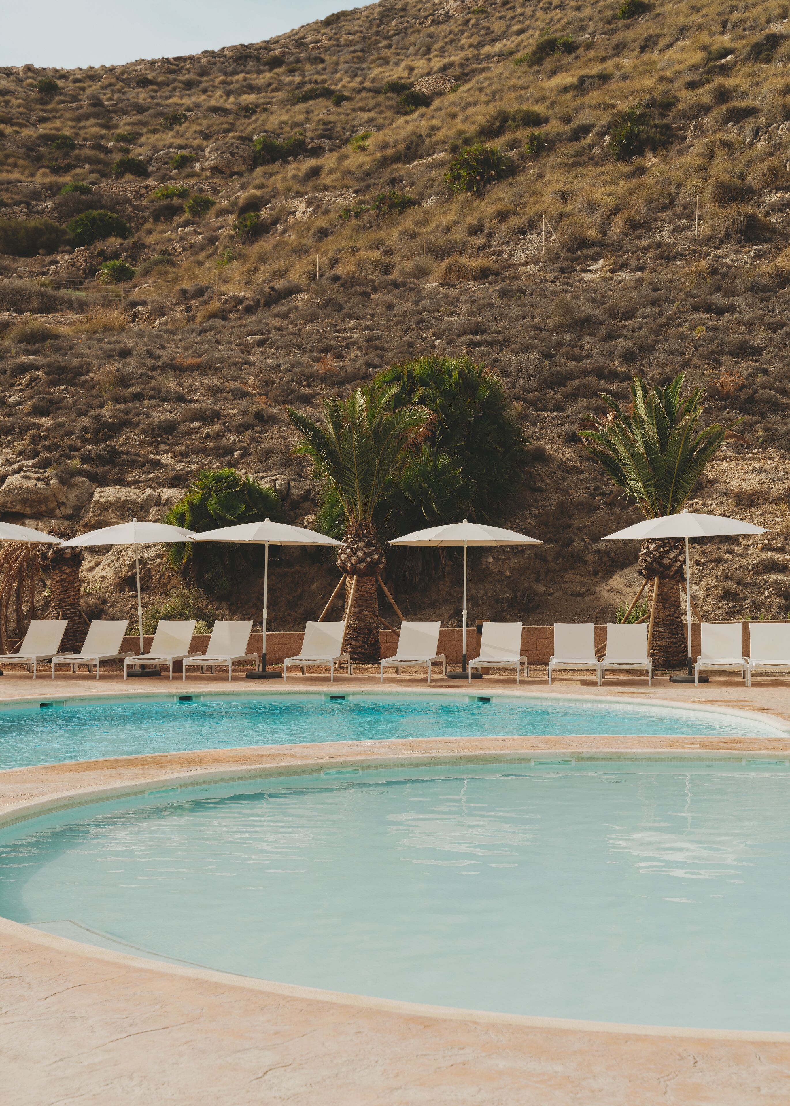 Zona de piscina con sillones de descanso y sombrillas, rodeada por un paisaje montañoso.