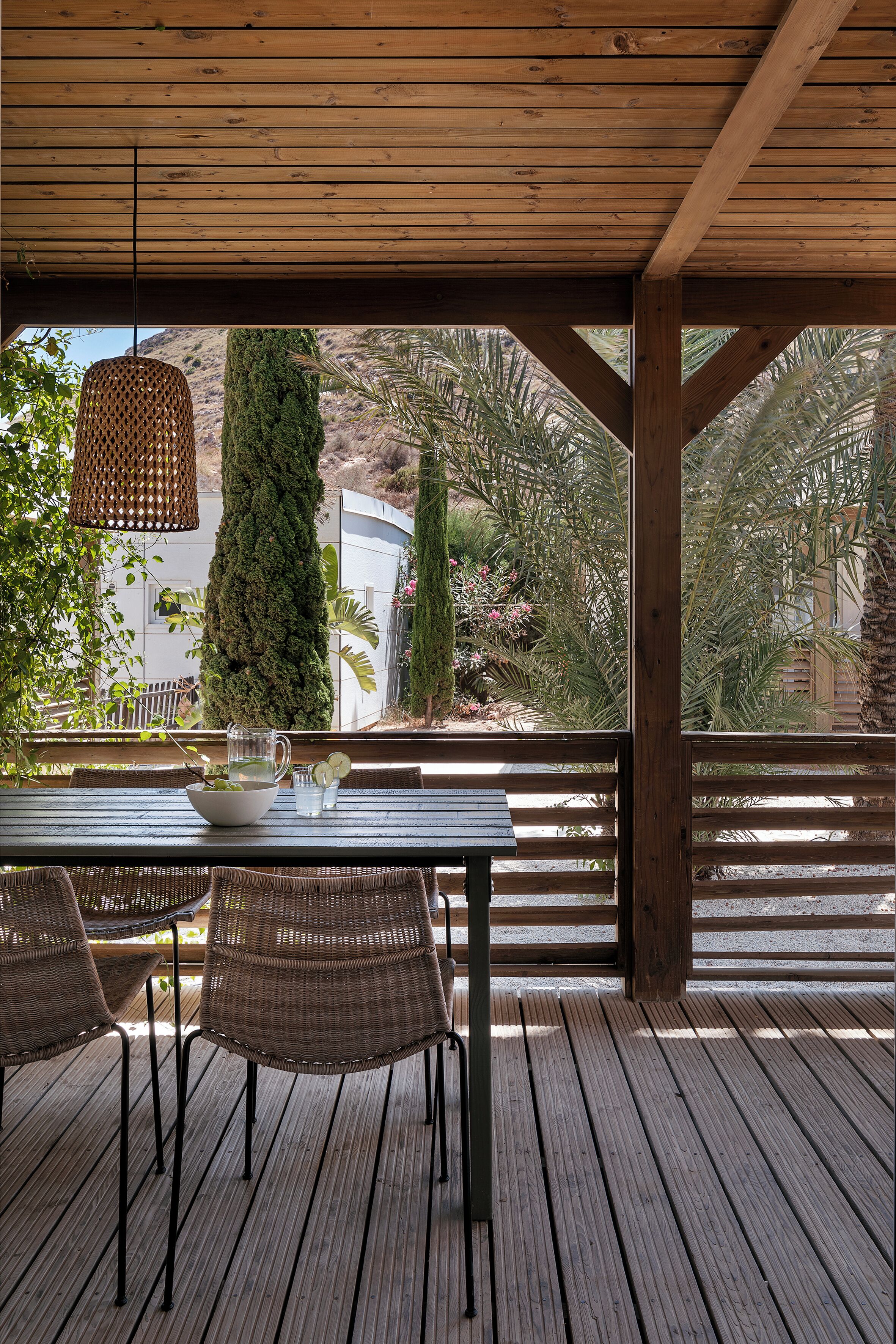 Una terraza de madera al aire libre con una mesa y sillas, rodeada de abundante vegetación y un árbol alto.