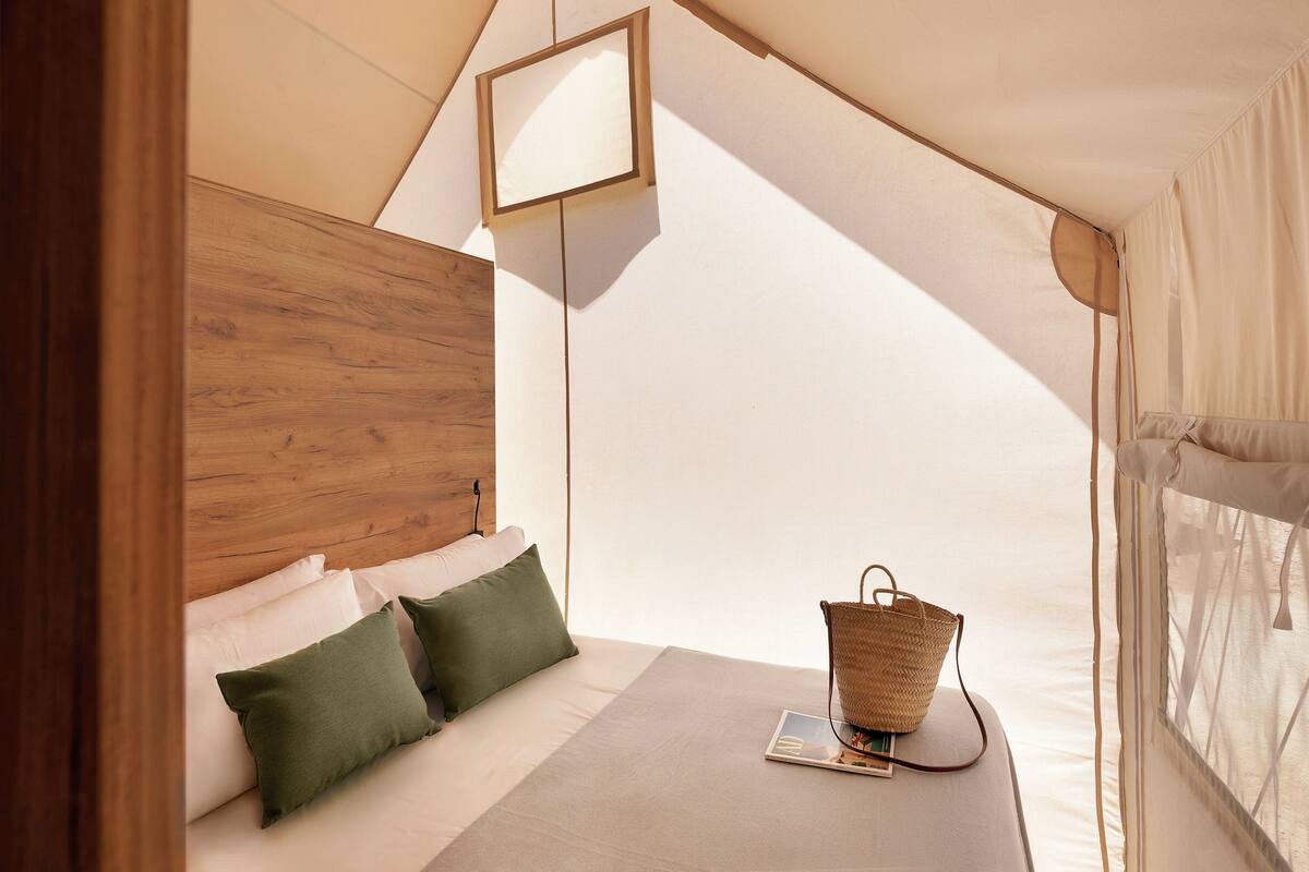 A tent-style room with a wooden headboard, a bed with green and white pillows, a small table with a basket, and a magazine.