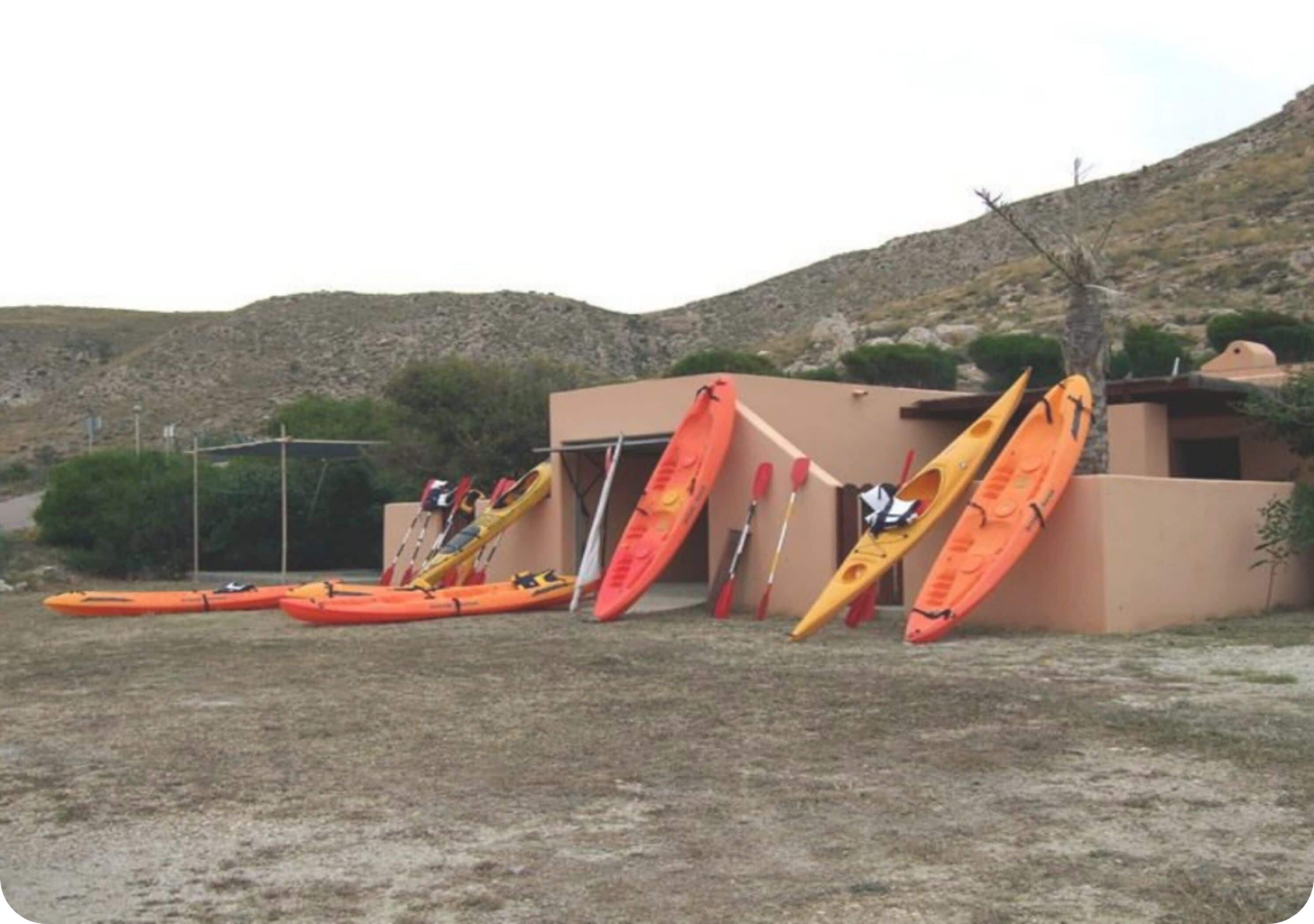 A row of kayaks leaning against a building in a dry, grassy area with hills in the background.