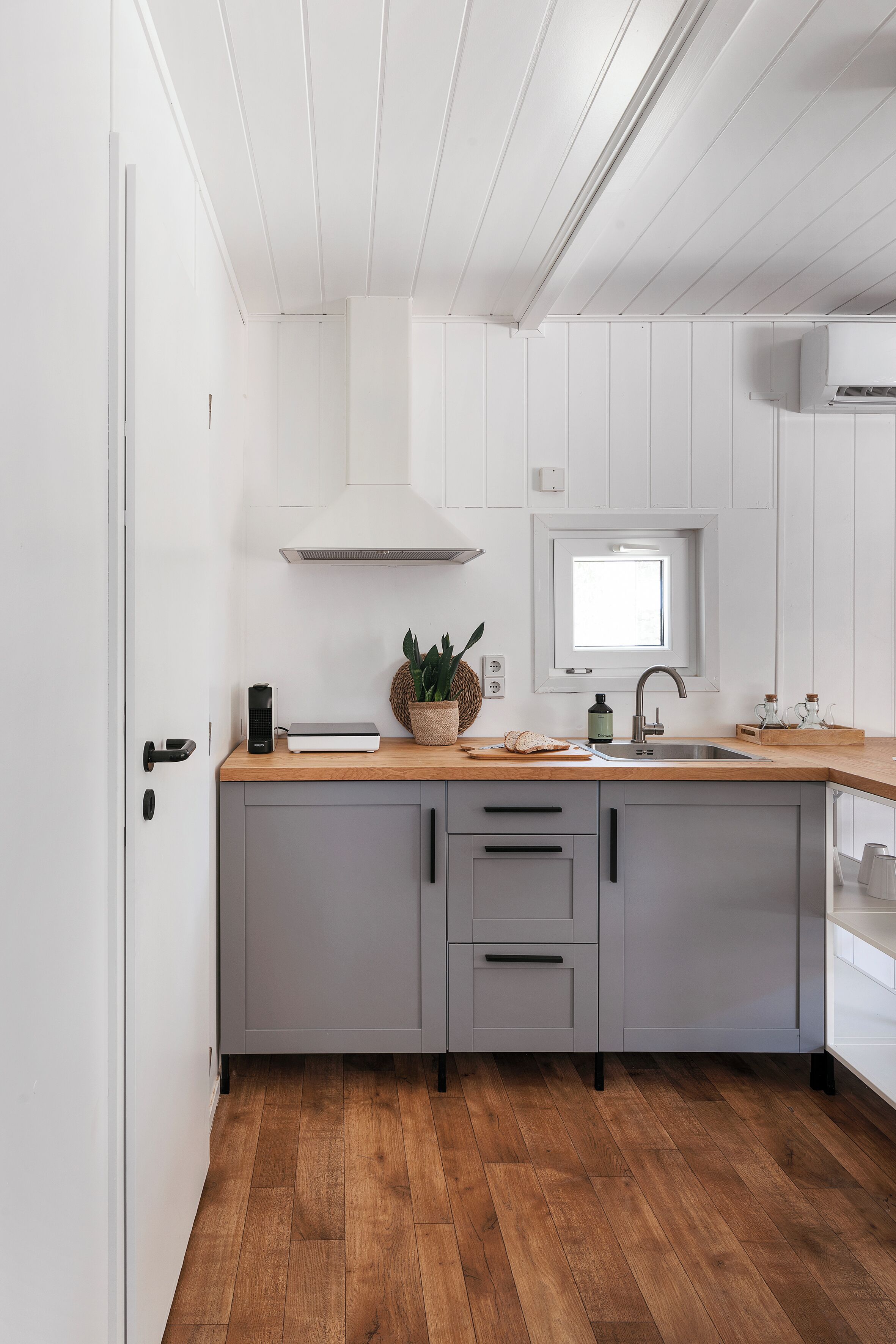 Una cocina moderna con encimera de madera, armarios grises, un ventanal sobre el fregadero y una puerta blanca.