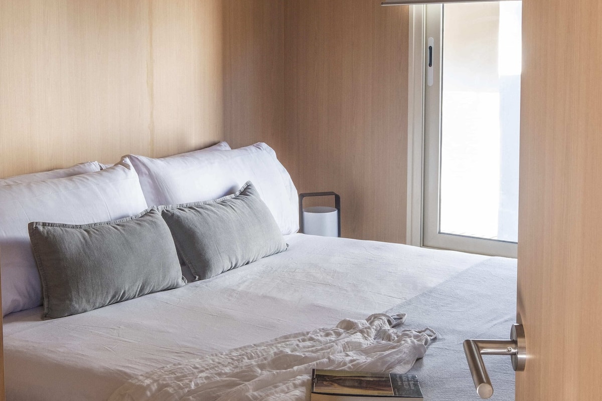 A neatly made bed with white and gray bedding, a bedside table with a book and a calculator, and a window with a white blind.