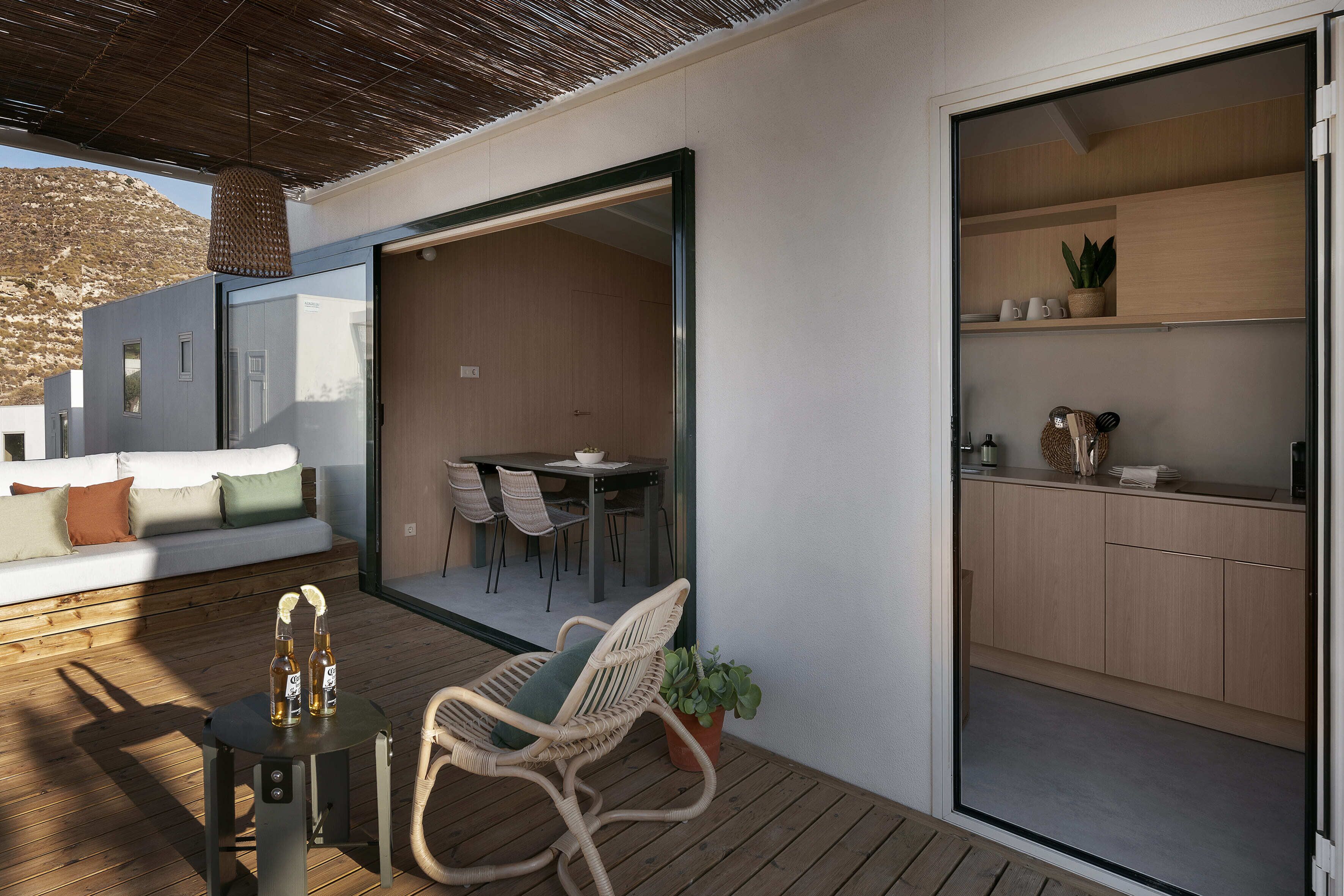 A modern outdoor living area with a wooden deck, a seating area with a sofa and chairs, and a sliding glass door leading to an interior space with a dining table and chairs.