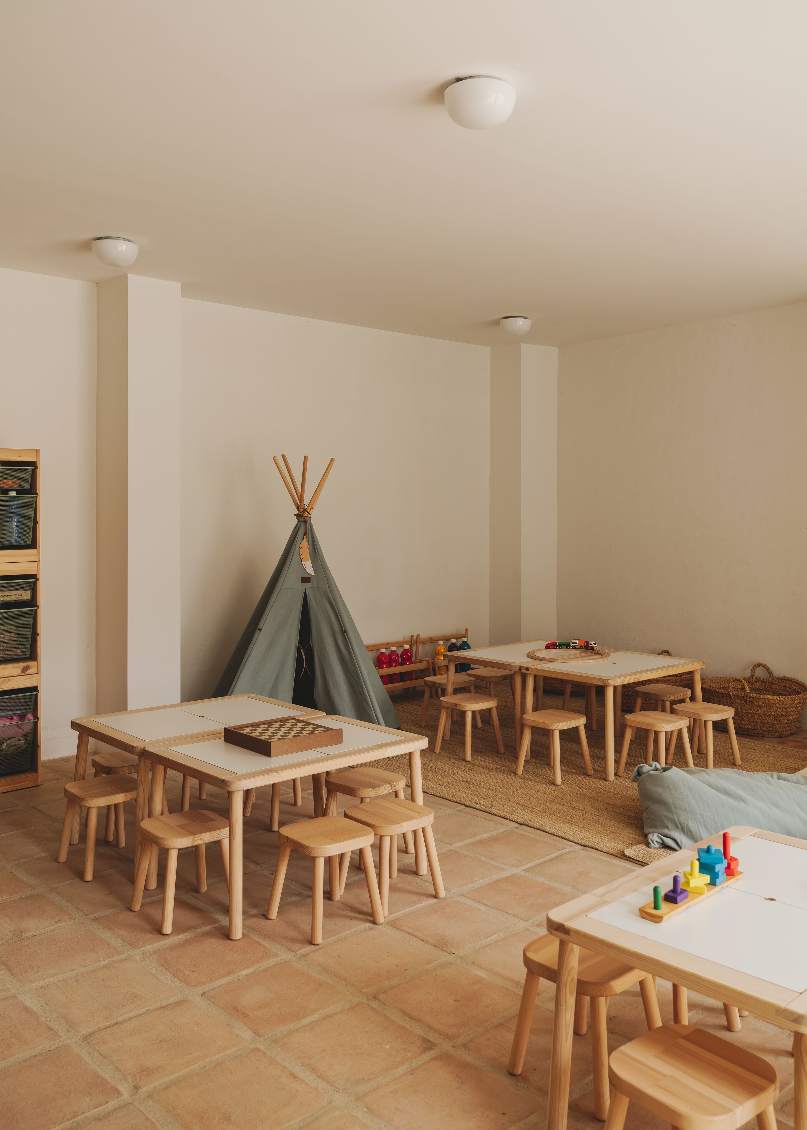 Une salle conçue pour les enfants, avec plusieurs tables et chaises en bois, une tente indienne verte et un espace de jeux équipé de jouets.