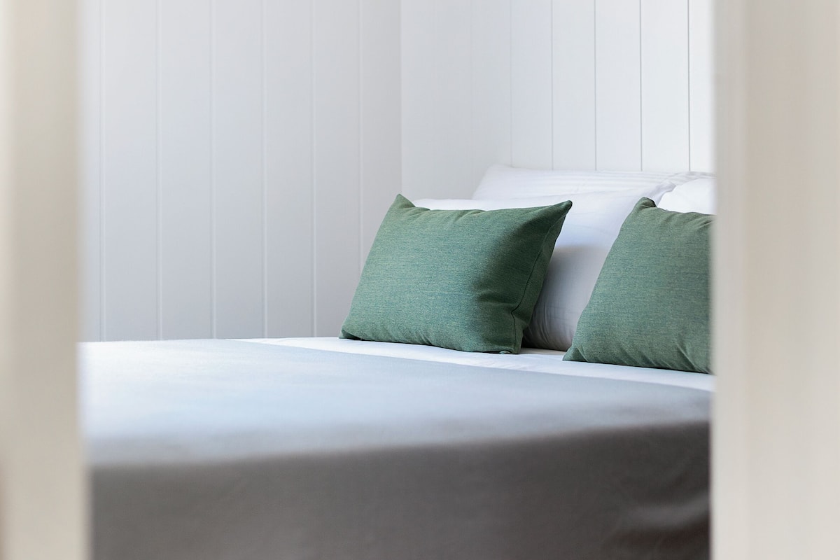 A neatly made bed with a white bedspread and two green pillows in a room with white paneled walls.