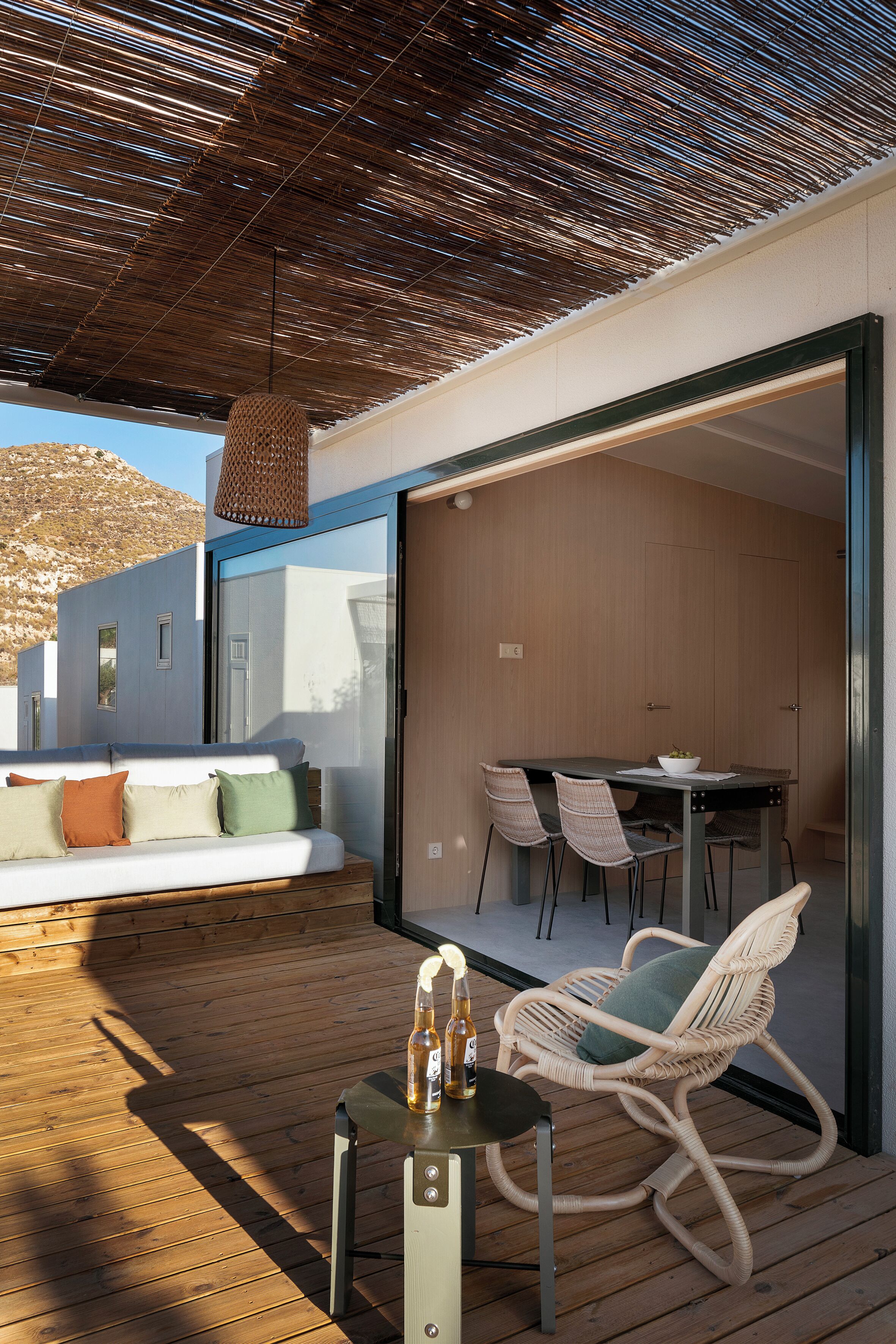 An outdoor seating area with a wooden deck, a wicker chair, a table with a bottle, and a glass partition wall.