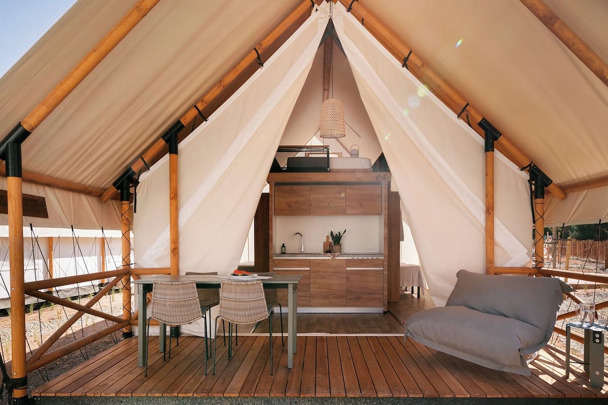 A tent with a wooden frame and a central sleeping area. There is a table and chairs outside the tent.