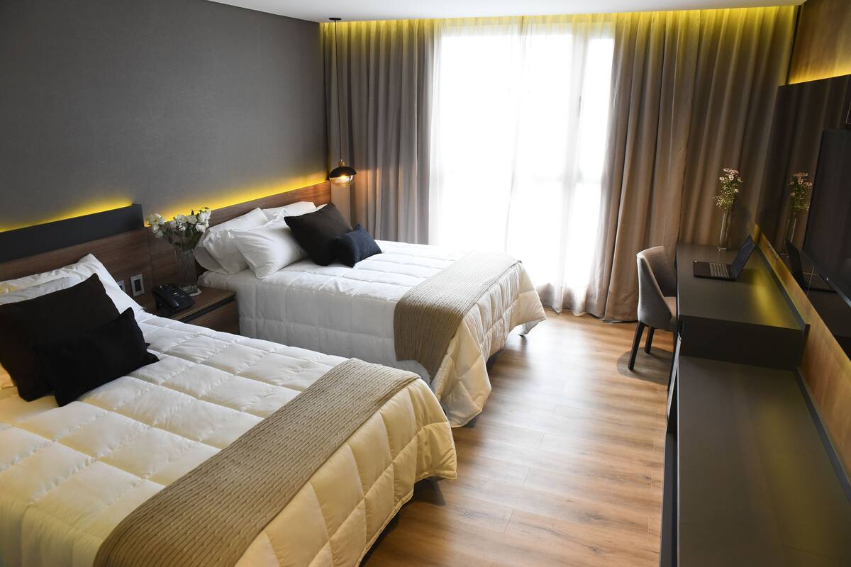 Ett hotellrum med två sängar, ett skrivbord, en stol och ett stort fönster med gardiner.