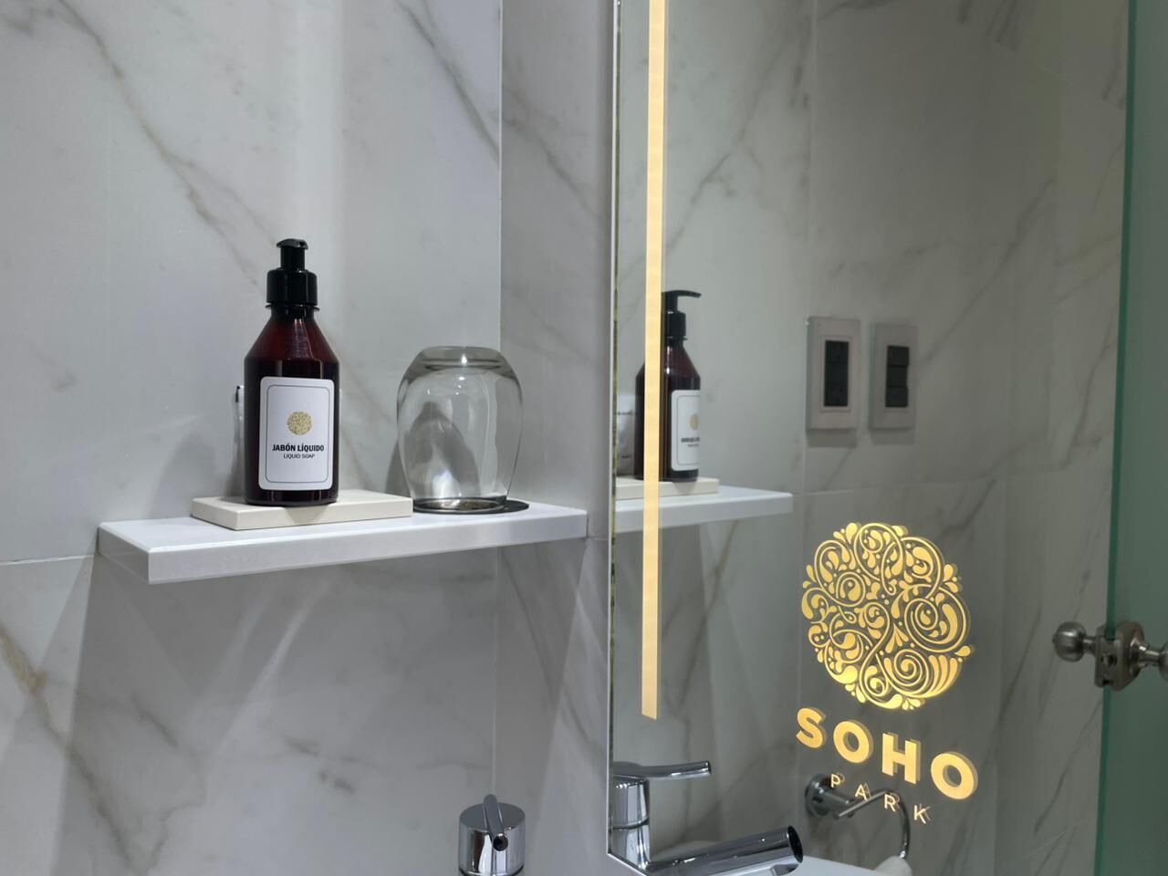 A bathroom with a marble wall, a white shelf with toiletries, a glass shower enclosure, and a door with a logo.