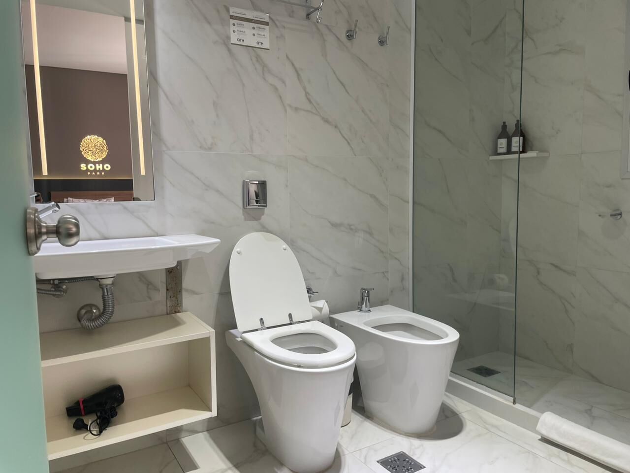 A bathroom with a marble wall, a sink, two toilets, a shower, and a mirror.