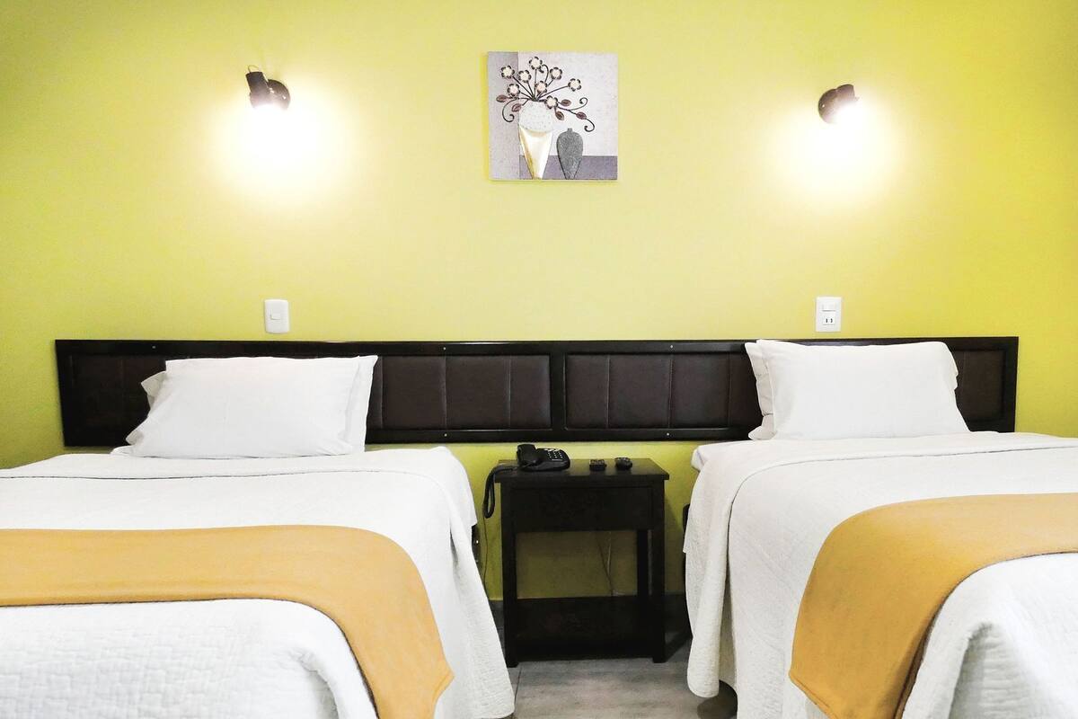 Une chambre d’hôtel avec deux lits, une table de chevet avec un téléphone et des télécommandes, et un luminaire fixé au mur.