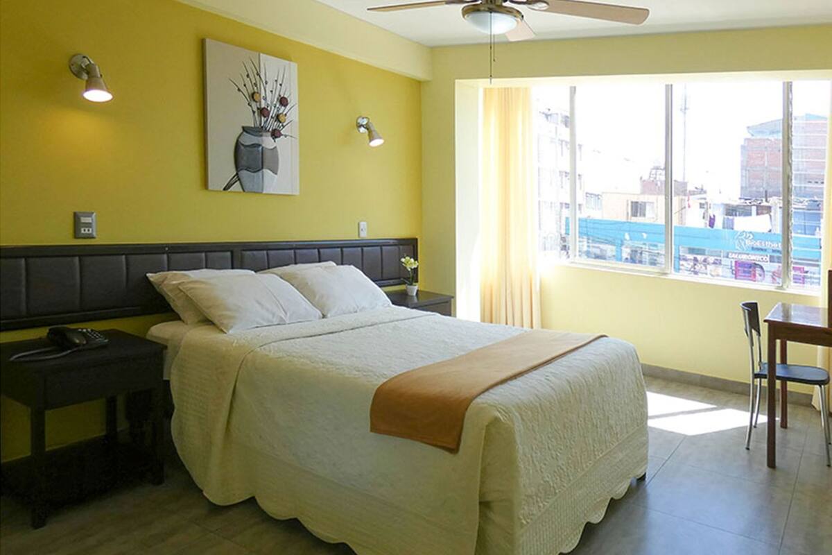 Une chambre d’hôtel équipée d’un lit, d’une table de chevet, d’une chaise, d’un ventilateur de plafond et d’une fenêtre donnant sur la ville.