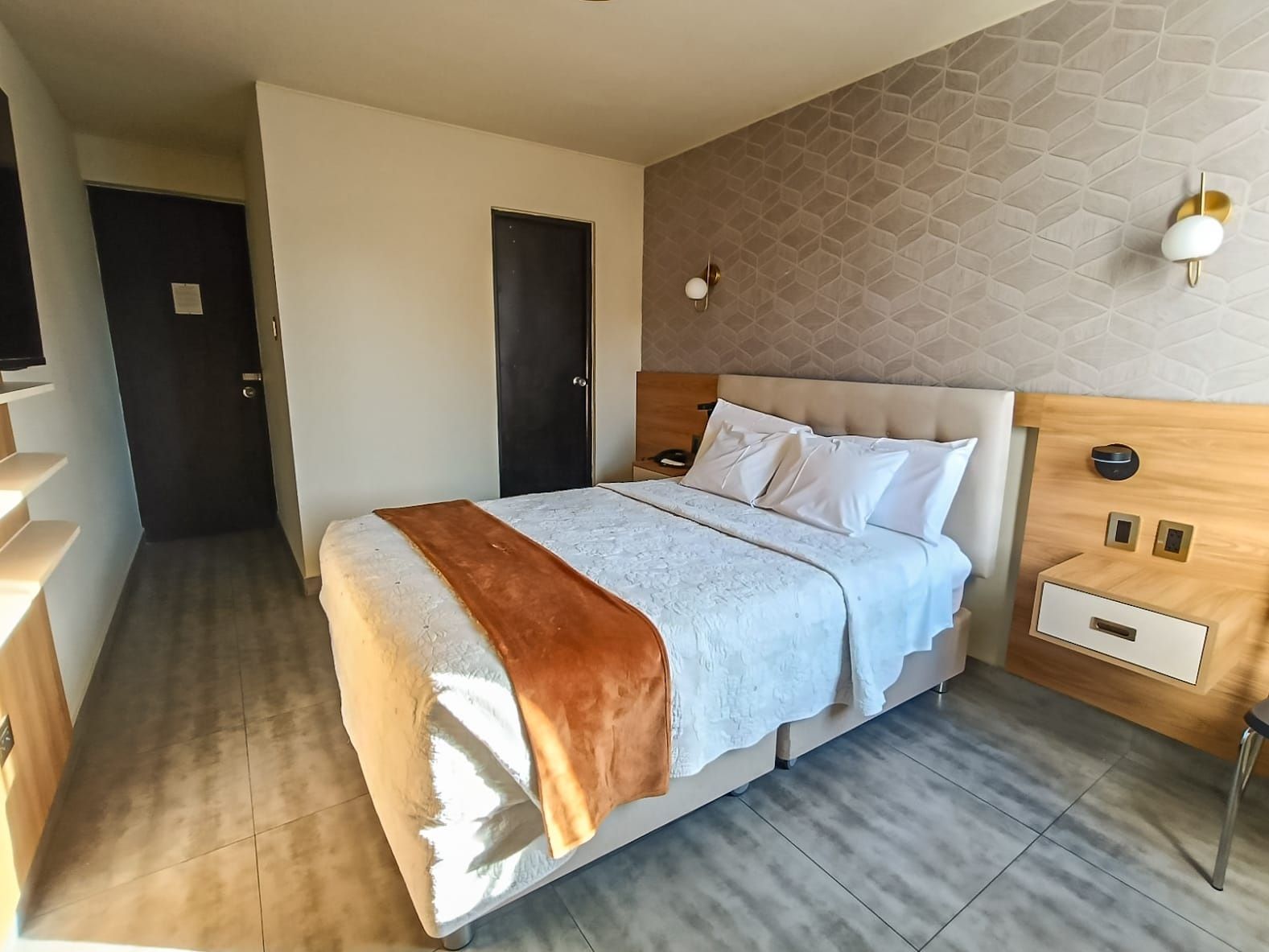 A hotel room with a bed, bedside tables, a wall-mounted light, and a door.