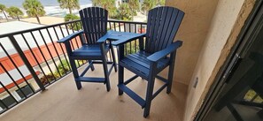Een balkon met twee Adirondack-stoelen en een kleine tafel, met uitzicht op een strand en palmbomen.