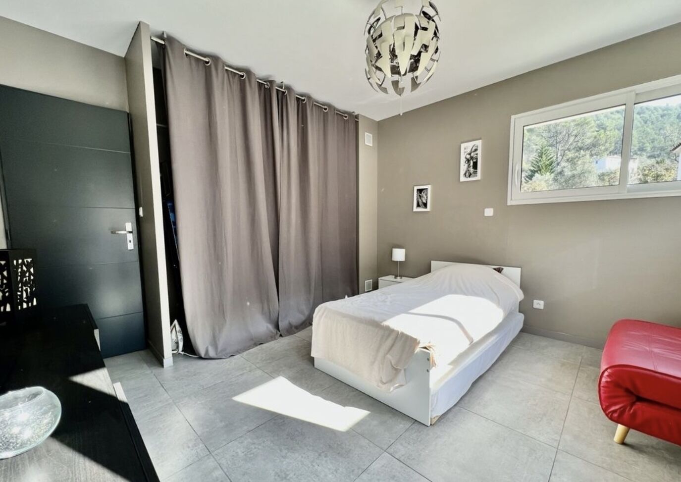 Un dormitorio moderno con una cama, un banco rojo, una puerta gris y una ventana con vistas a árboles.