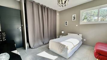 Un dormitorio moderno con una cama, un banco rojo, una puerta gris y una ventana con vistas a árboles.