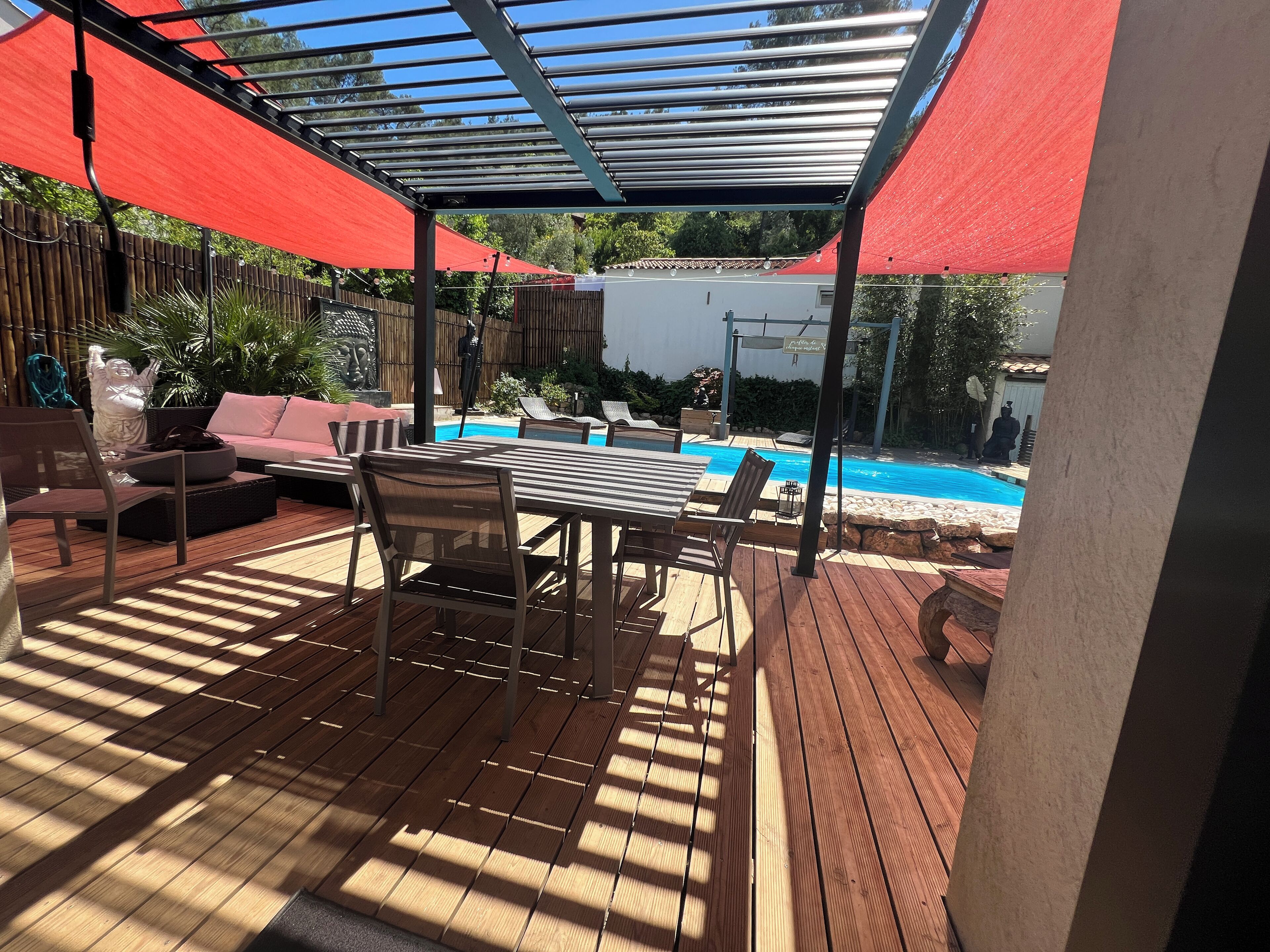 An outdoor patio with a pool, wooden deck, and red umbrellas providing shade.