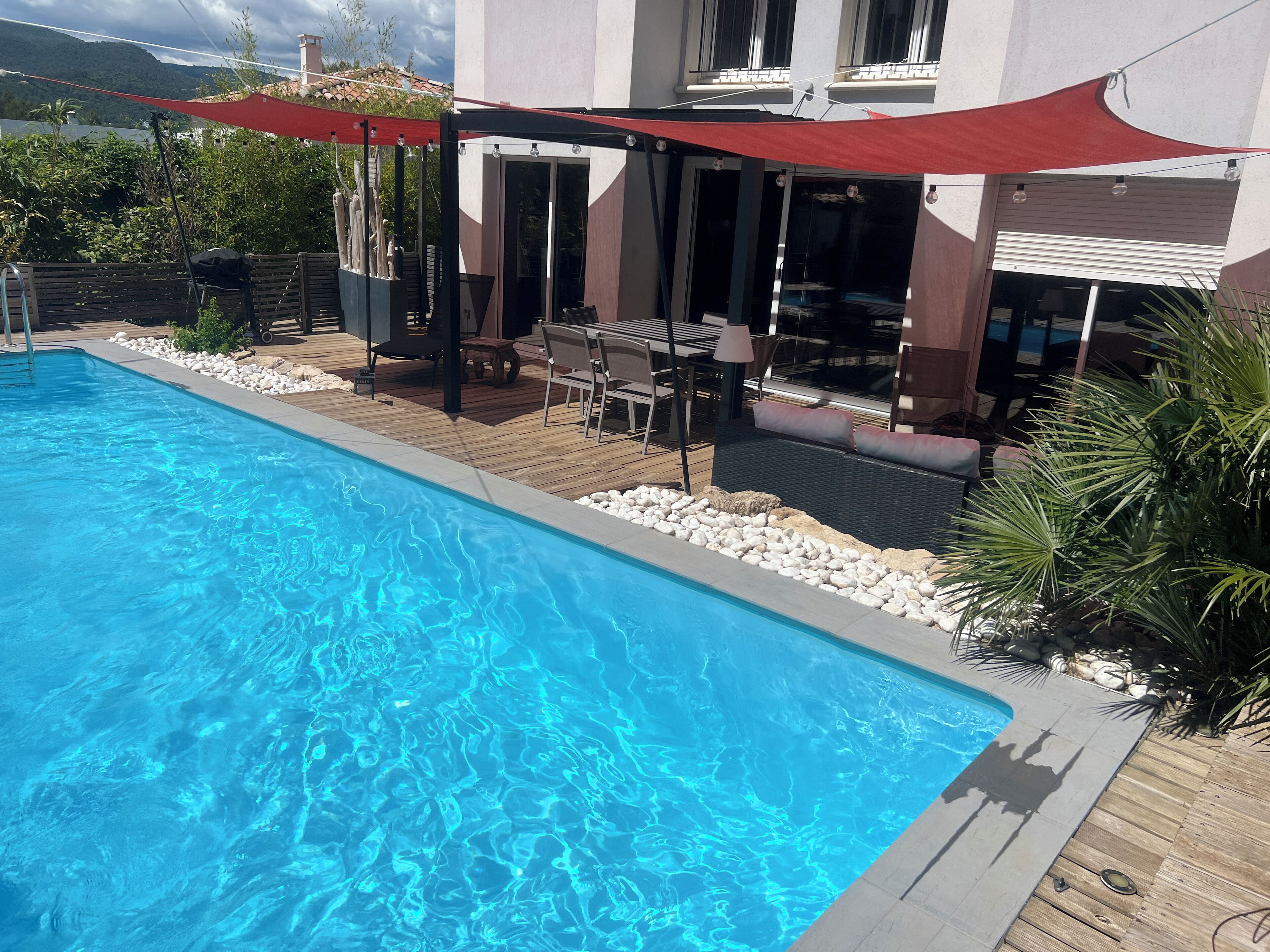 Une piscine avec une terrasse en bois, des sièges d’extérieur et un auvent rouge.