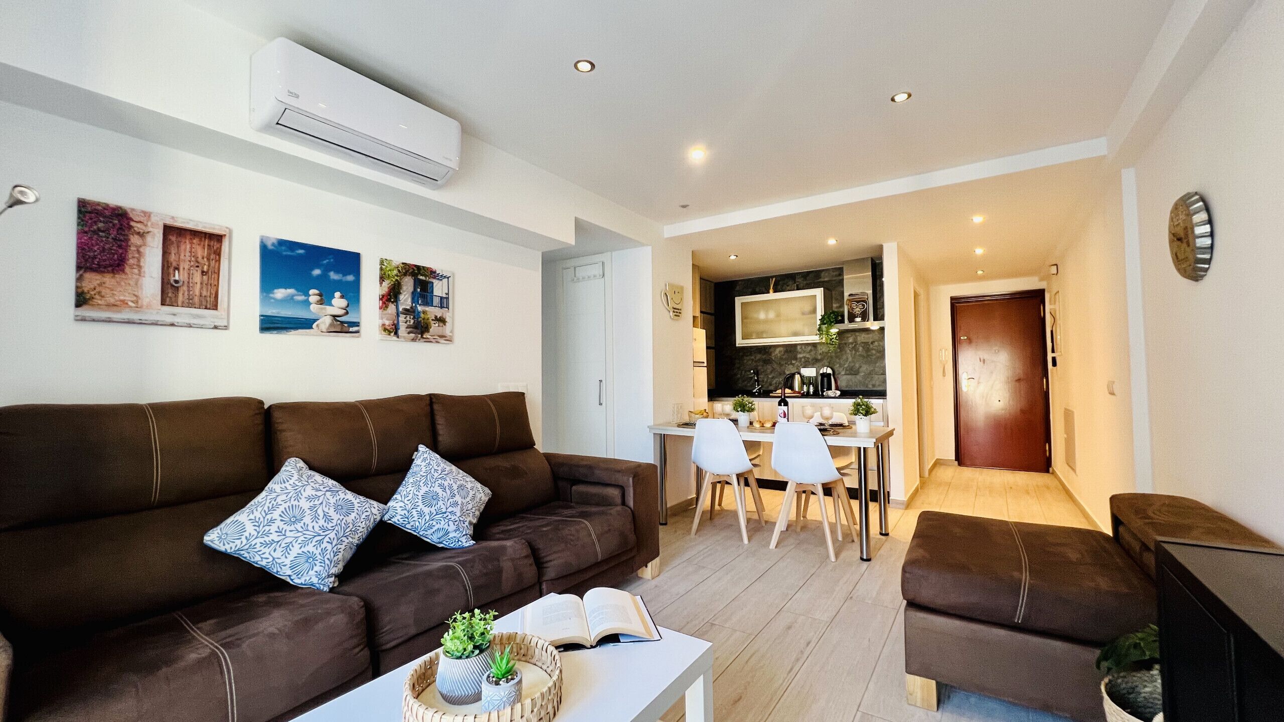 A modern living room with a brown sofa, a dining area with a white table and chairs, and a kitchen in the background.