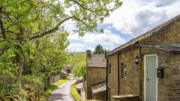 A narrow stone pathway leading to a traditional stone cottage with a light blue door, surrounded by lush greenery and a stone wall.