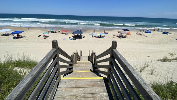 Ein hölzerner Steg führt zu einem Strand mit Sonnenschirmen und Menschen.