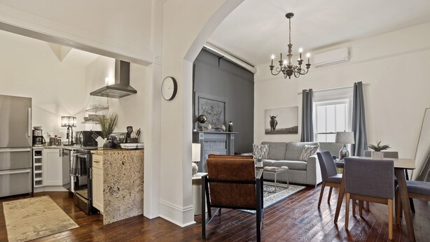 A modern kitchen with a central island, a dining area with a chandelier, and a living room with a sofa and chairs.