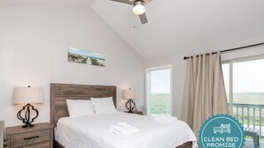 A clean, well-lit bedroom with a wooden headboard, white bedding, and a view of the outdoors through large windows.