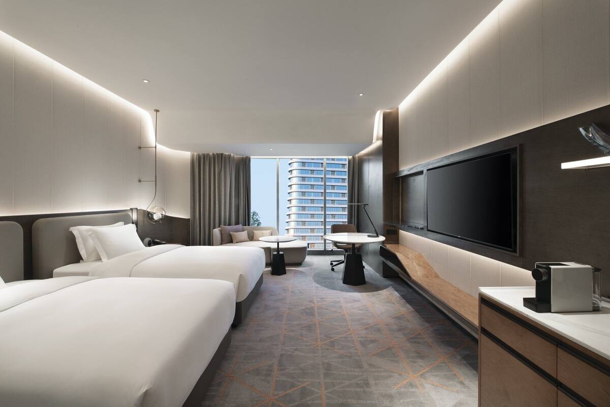 Ein Hotelzimmer mit zwei Betten, einem Schreibtisch, einem Fernseher und einem großen Fenster mit Blick auf ein Hochhaus.