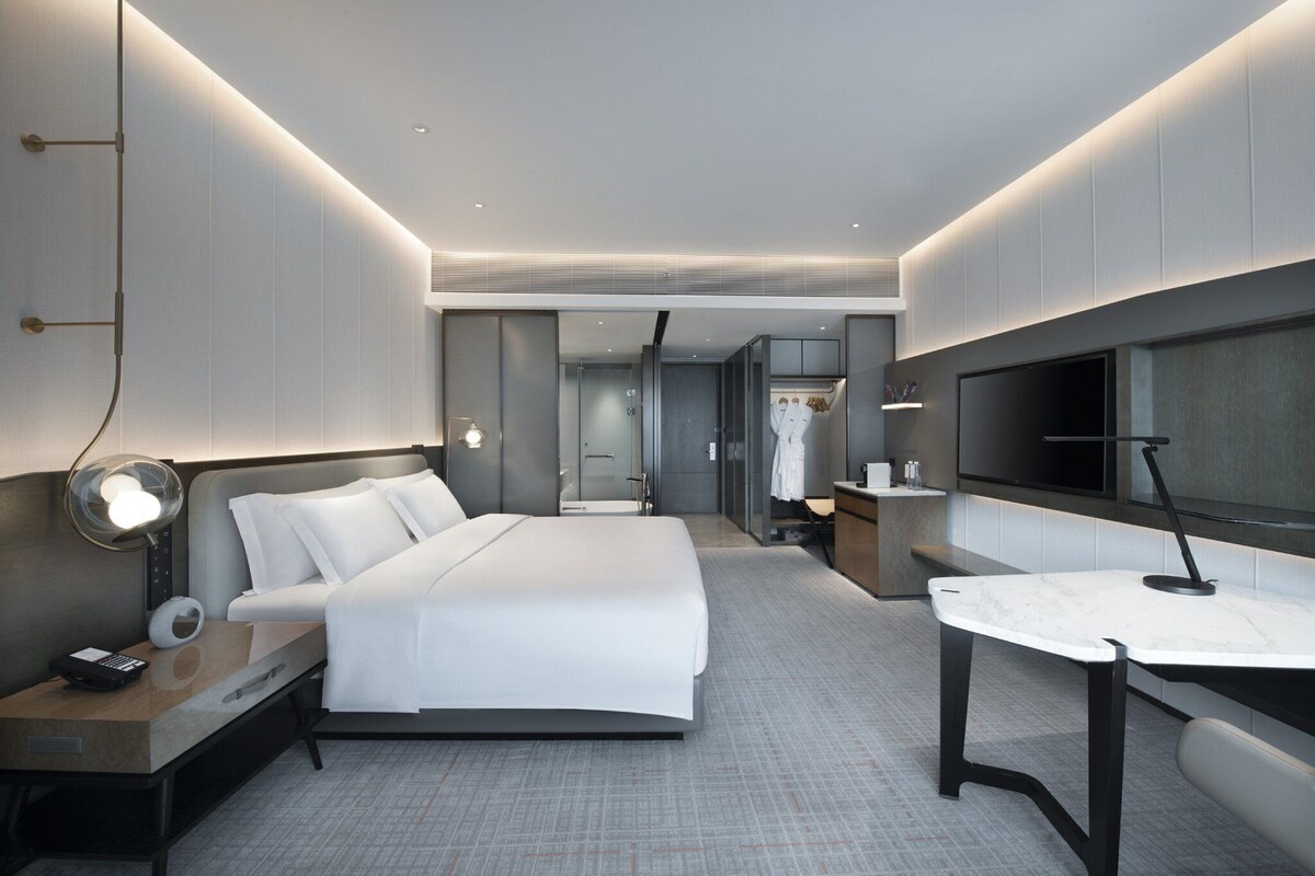 A modern hotel room with a large bed, bedside table with a lamp, a small desk with a chair, a TV, and a walk-in closet.