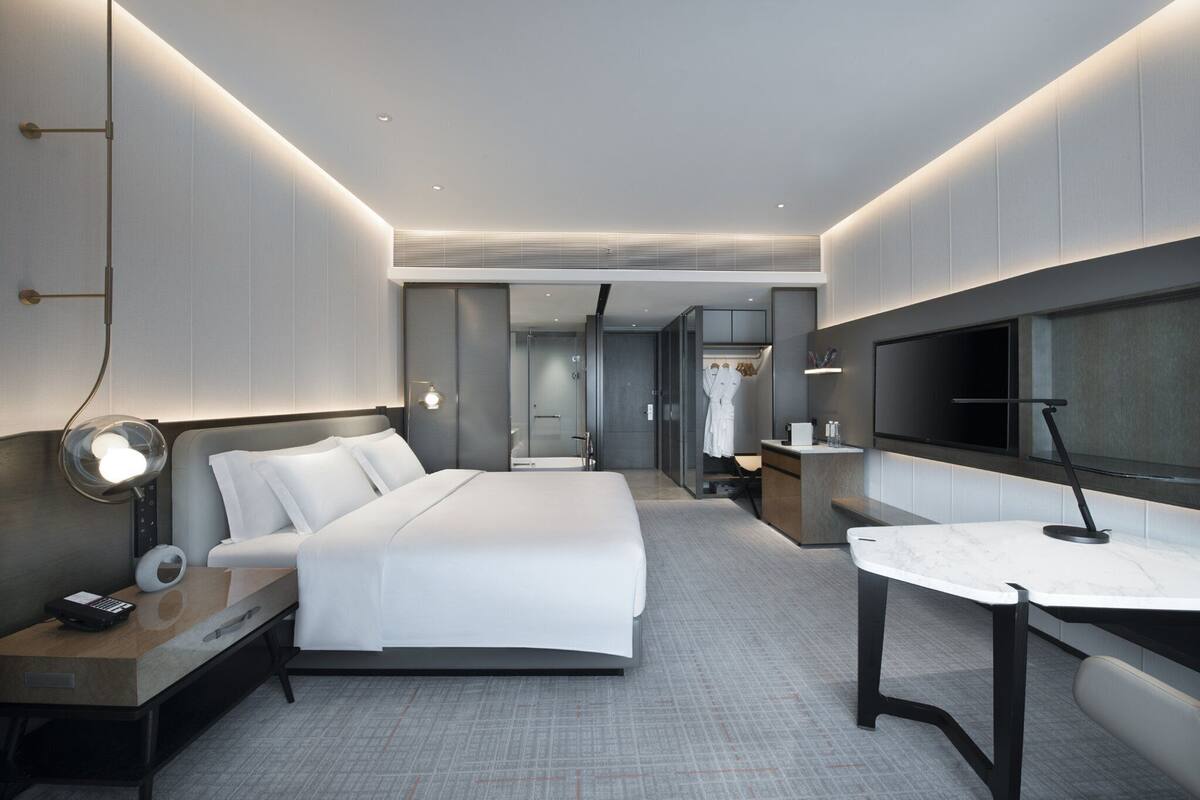 Ein modernes Hotelzimmer mit einem großen Bett, einem Nachttisch mit Lampe, einem kleinen Schreibtisch mit Stuhl, einem Fernseher und einem begehbaren Kleiderschrank.
