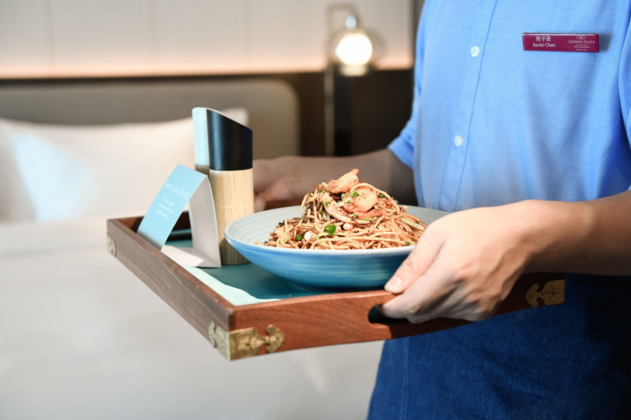 A person in a blue shirt carrying a tray with a blue plate of noodles and a wooden knife block.
