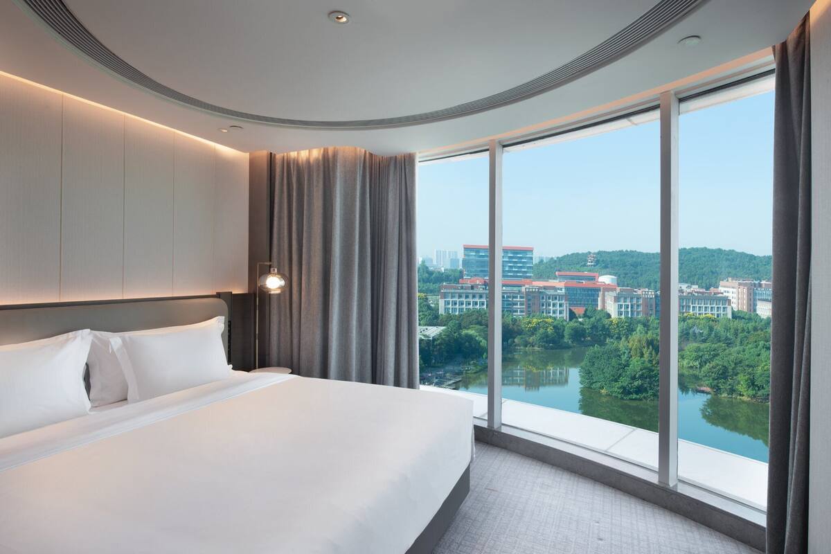 Ein modernes Hotelzimmer mit einem großen Bett, Blick auf einen Fluss und Gebäude sowie einem großen Fenster mit Vorhängen.