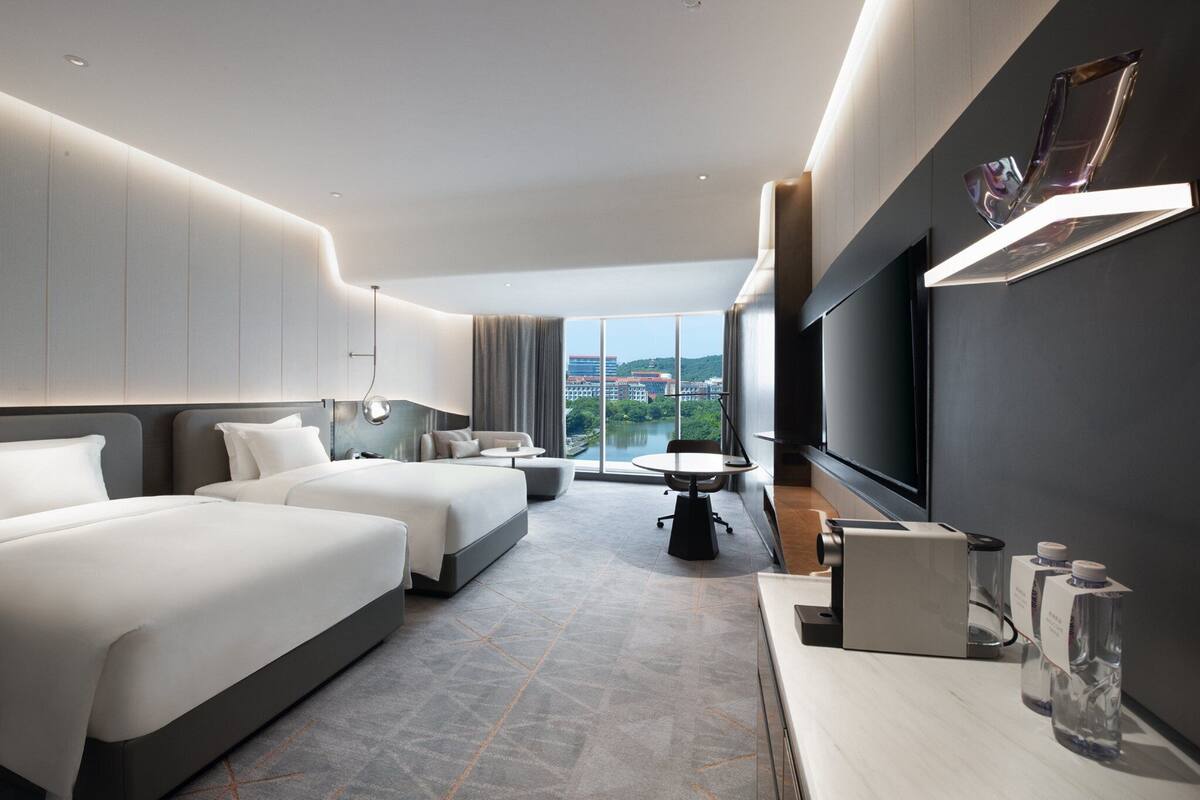 Ein modernes Hotelzimmer mit zwei Betten, einem Schreibtisch, einem Flachbildfernseher und einem großen Fenster mit Blick auf die Stadt.