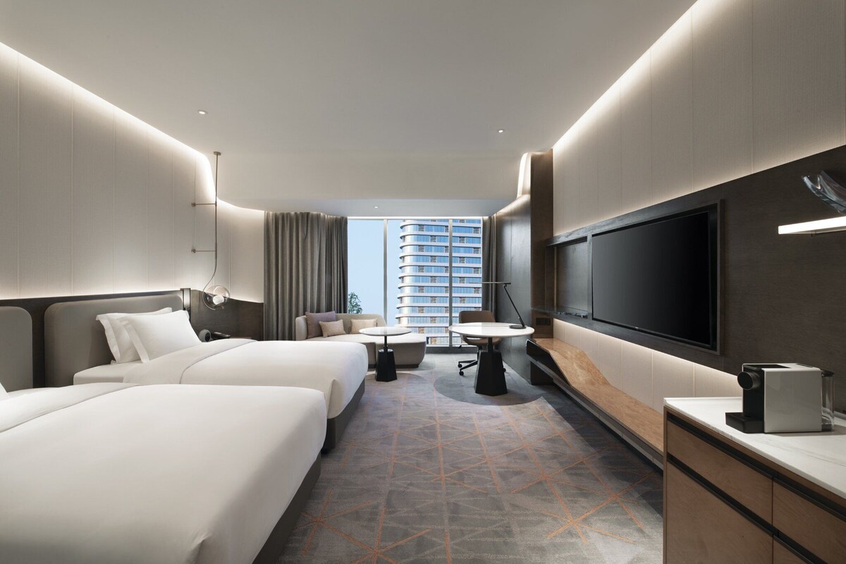 A hotel room with two beds, a desk, a TV, and a large window with a view of a high-rise building.