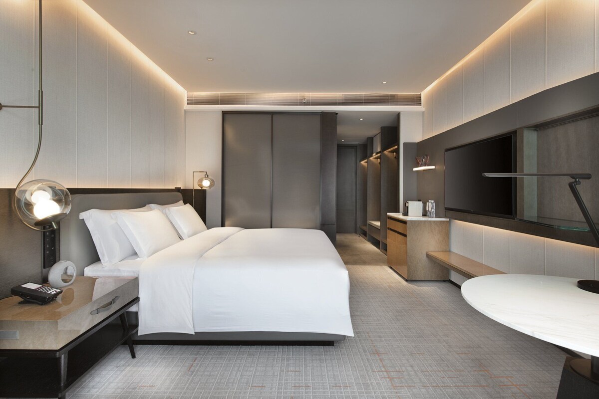 A modern hotel room with a large bed, bedside tables, a desk, a flat-screen TV, and a large mirror.