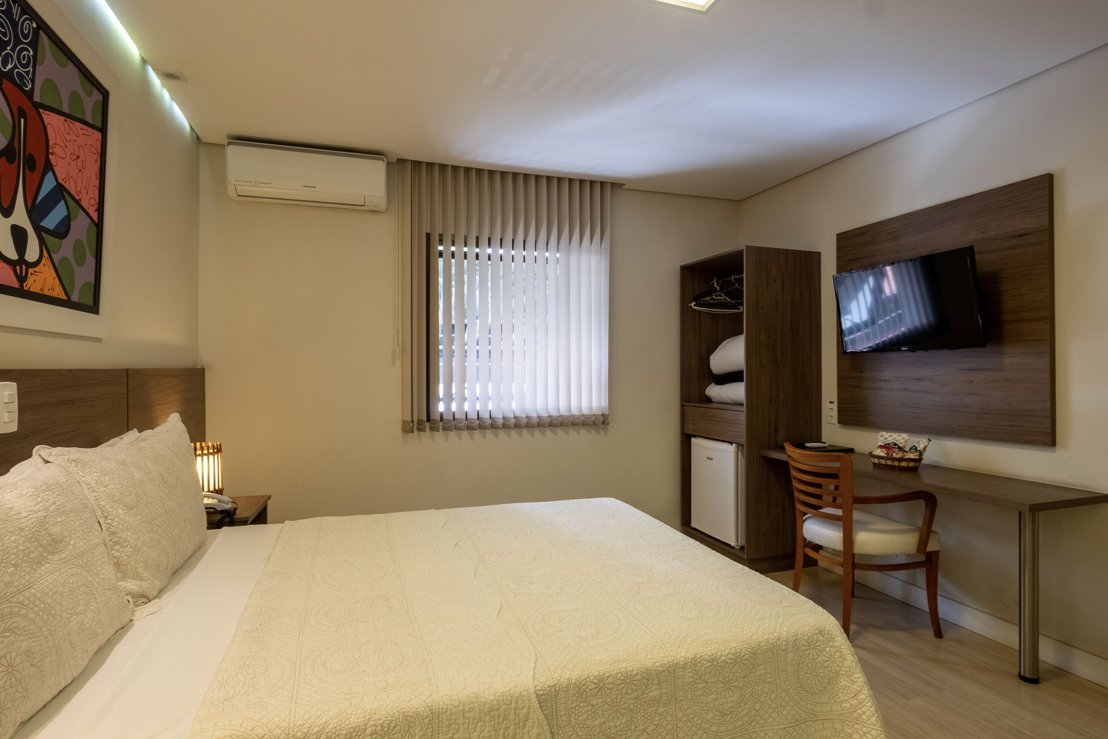 A hotel room with a bed, a desk with a chair, a television mounted on the wall, a wardrobe, and a window with blinds.