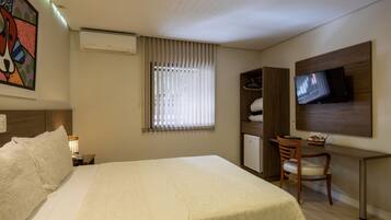 A hotel room with a bed, a desk with a chair, a television mounted on the wall, a wardrobe, and a window with blinds.