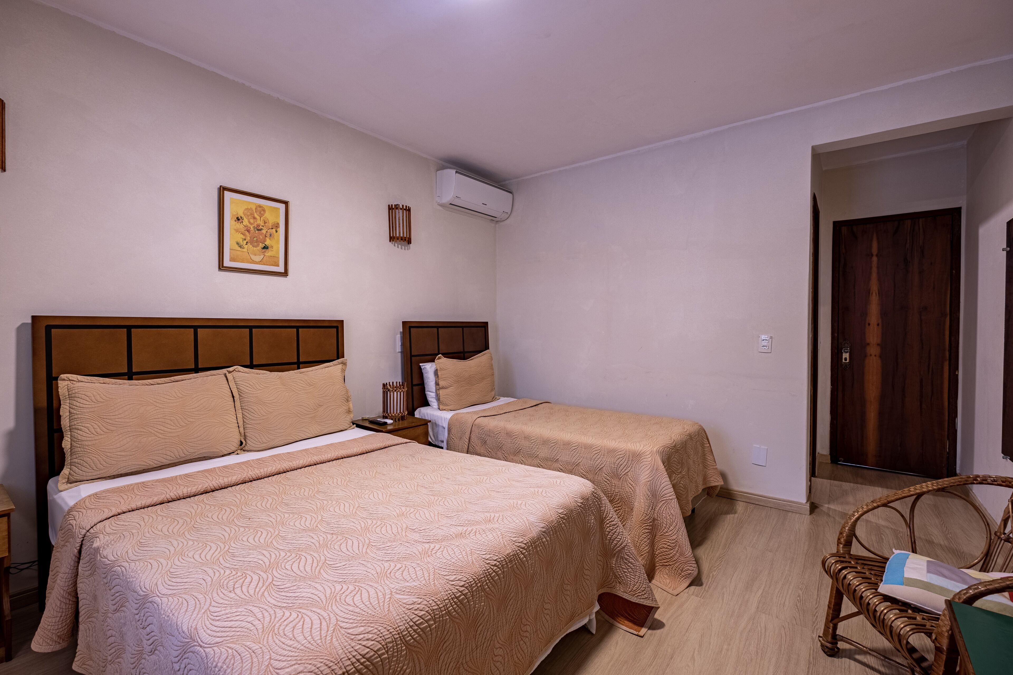 Quarto de hotel com duas camas, cabeceira de madeira, ar condicionado fixado na parede e um quadro emoldurado.