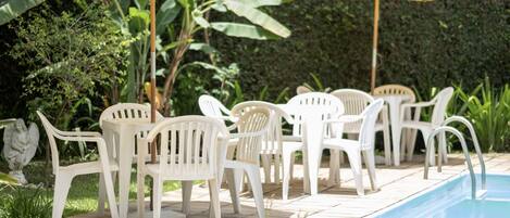 A poolside area with white plastic chairs and two umbrellas, surrounded by greenery.
