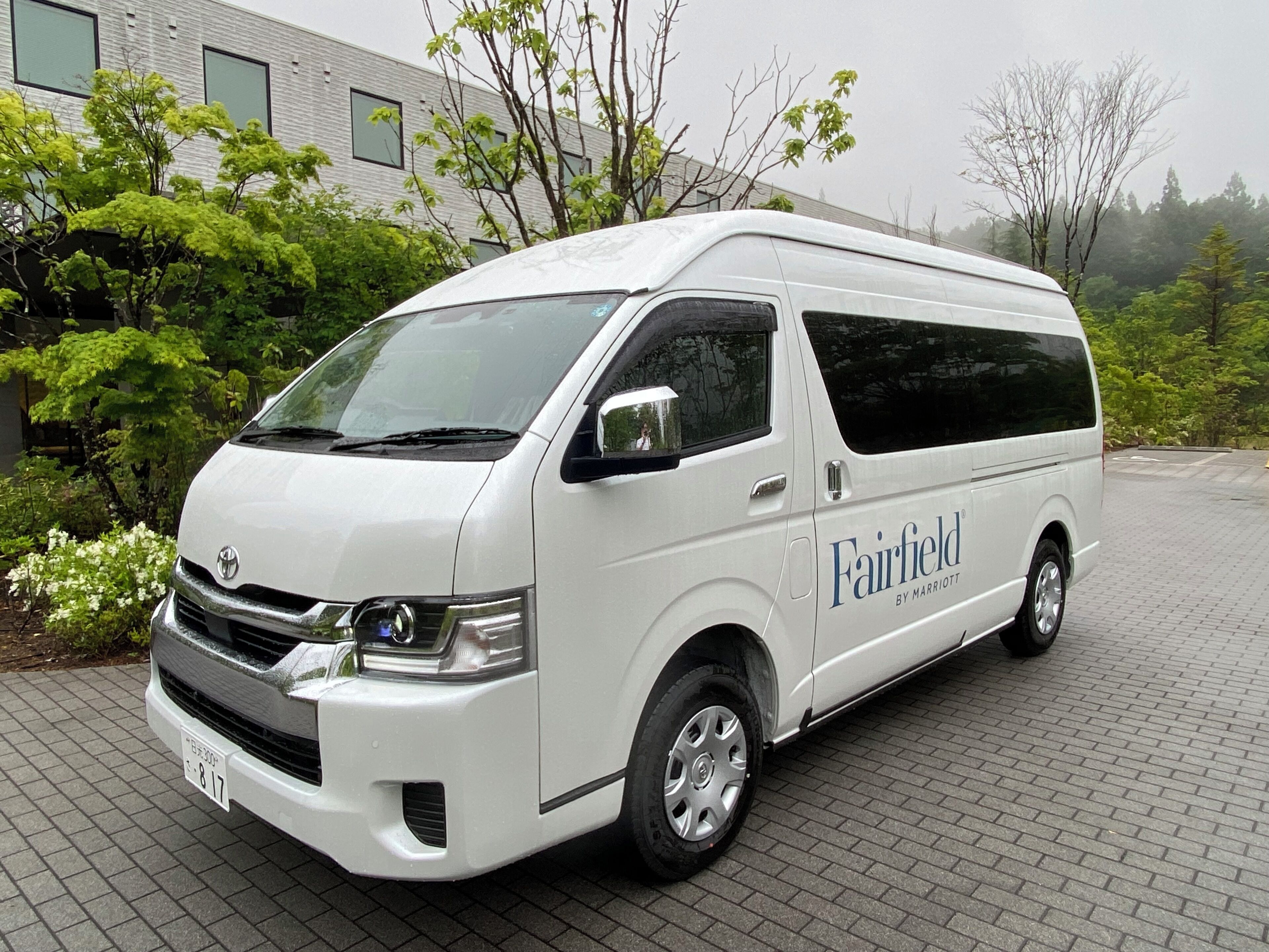 A white van with 'Fairfield by Marriott' branding parked on a paved area with greenery and a building in the background.
