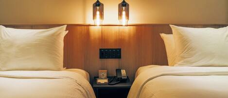Habitación de hotel con dos camas, un cabecero de madera, una mesita de noche con teléfono y reloj, y dos lámparas colgantes.
