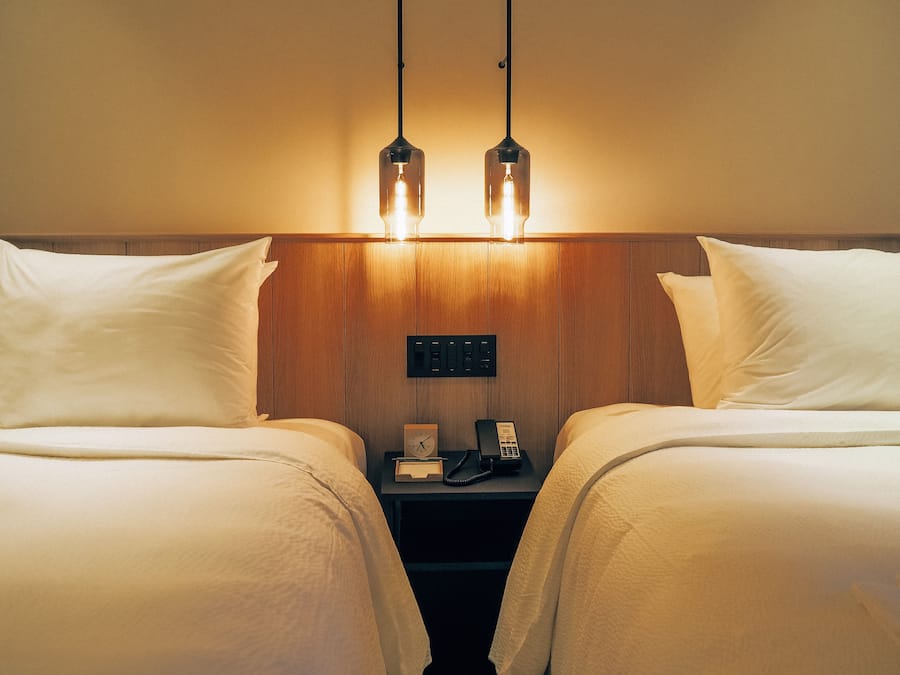 Habitación de hotel con dos camas, un cabecero de madera, una mesita de noche con teléfono y reloj, y dos lámparas colgantes.