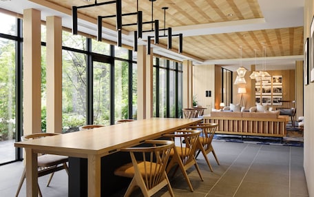 A spacious dining area with a long wooden table and chairs, large windows offering a view of greenery, and a cozy seating area with a sofa and lamps.