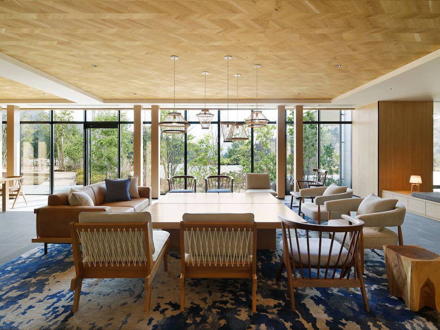 A spacious room with a large wooden table surrounded by chairs, a sofa, and a coffee table. Large windows provide natural light and views of greenery outside.
