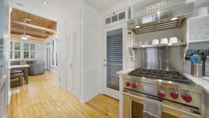 Ett modernt kök med trägolv, vita köksluckor, en spis i rostfritt stål och en matplats med en grå soffa.